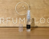 A 2ml spray bottle with a black label and transparent cap is placed in front of a wooden background with the word "Perfumology" and a bird logo engraved on it, exuding an air of sophistication that hints at the woody white musk perfume Boutique within.