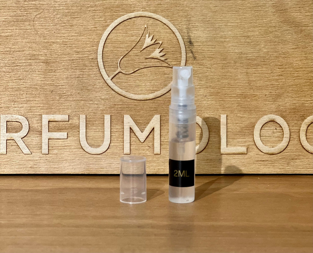 A 2ml spray bottle with a black label and transparent cap is placed in front of a wooden background with the word "Perfumology" and a bird logo engraved on it, exuding an air of sophistication that hints at the woody white musk perfume Boutique within.