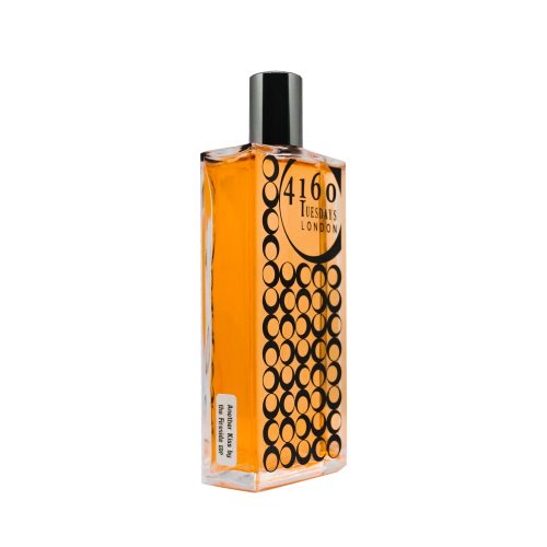 A rectangular perfume bottle with orange liquid, labeled "Another Kiss by the Fireside" and decorated with black circles, has a black cap. This floral fougere evokes a sophisticated perfume dialect from 4160Tuesdays.