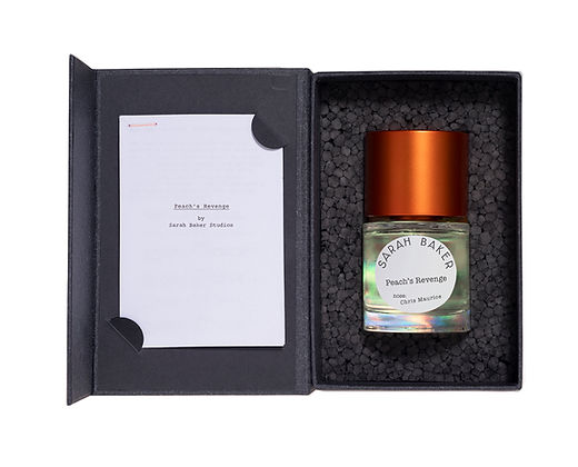 A perfume bottle labeled "Peaches Revenge" with a copper-colored cap sits in a black box beside a card titled "Peaches Revenge by Sarah Baker," emanating subtle hints of vanilla and spices.