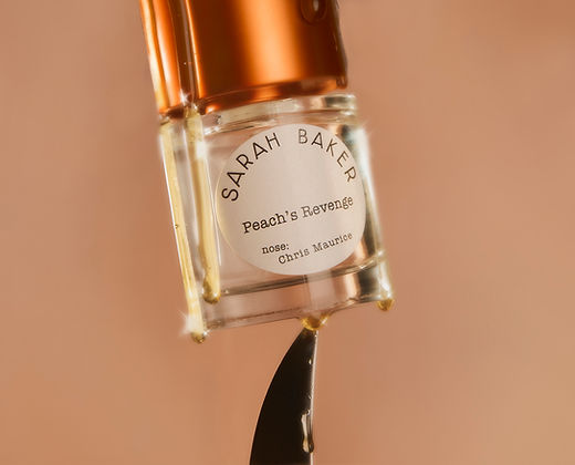 A close-up image of a perfume bottle labeled "Sarah Baker Peaches Revenge." The bottle is upside down, and some liquid is dripping from it. The label also mentions "nose: Chris Maurice." A hint of peach and spices subtly lingers around the scene.