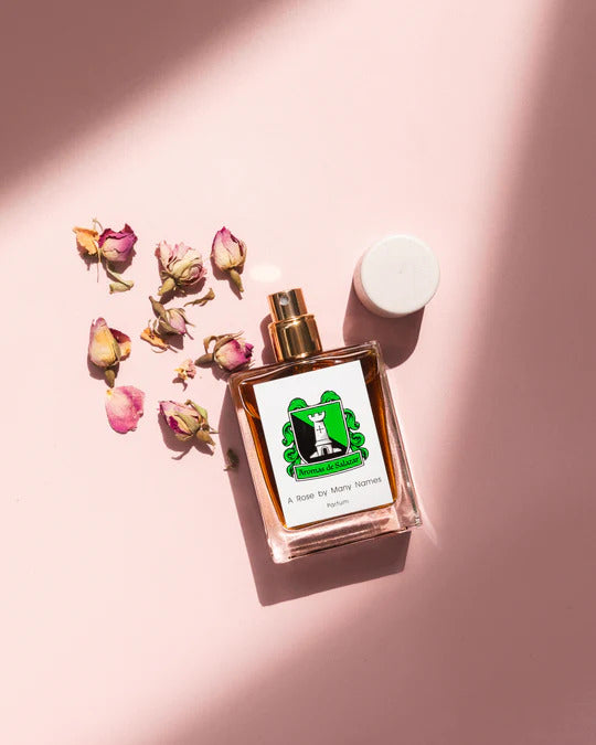 A square perfume bottle labeled "A Rose by Many Names" with pink liquid rests on a pink surface, surrounded by dried rose petals. The rose perfume from Aromas de Salazar features a white cap and a green logo with a lighthouse.
