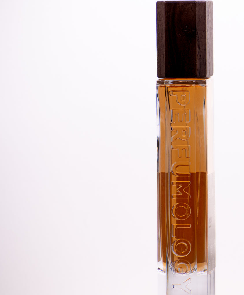 A rectangular glass perfume bottle with a wooden cap is filled with amber-colored liquid. The word "Perfumology" is embossed vertically on the side, showcasing their dedication to high quality ingredients. The background is plain white. The product name "Blyss" enhances the allure of this exquisite fragrance.