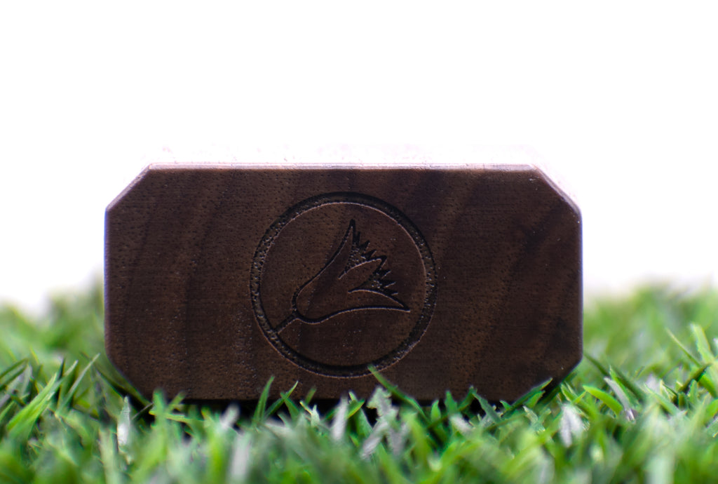 A wooden block with the Perfumology Blyss fragrance logo is placed on green grass, with a white background.