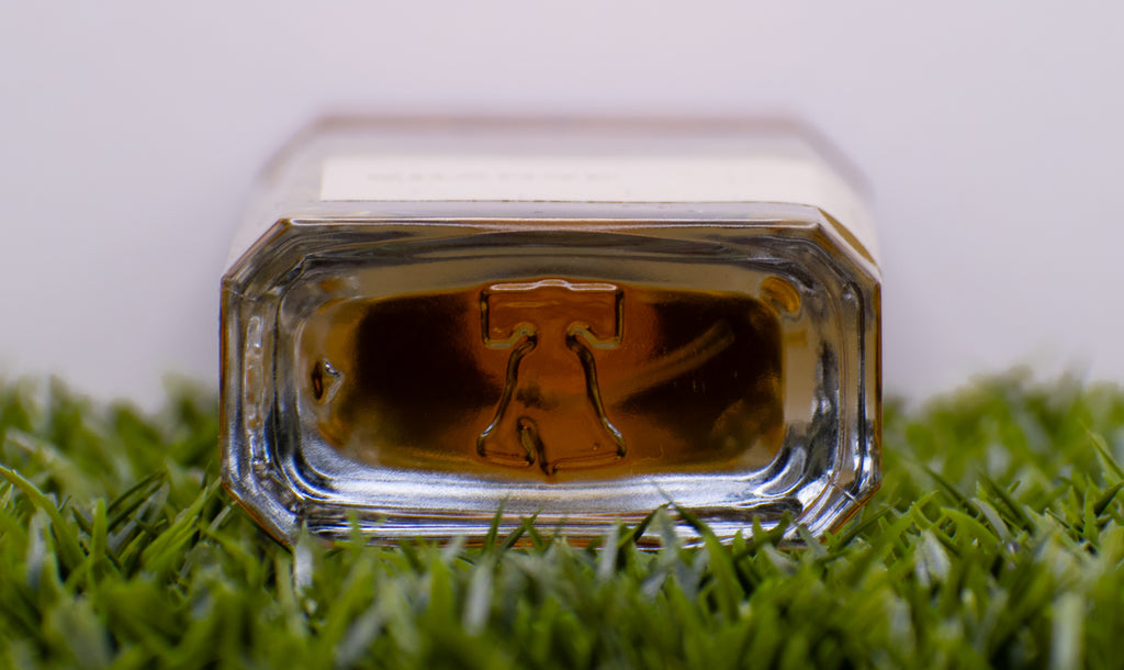 A close-up of the bottom of a hexagonal glass bottle featuring an embossed Liberty Bell design, resting on green artificial grass, encapsulating the essence of Blyss fragrance crafted by Perfumology with high quality ingredients.