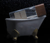 A clear glass perfume bottle with a wooden cap labeled "Boutique" by Perfumology, exuding hints of woody white musk, is elegantly placed on a decorative miniature porcelain bathtub with gold accents.