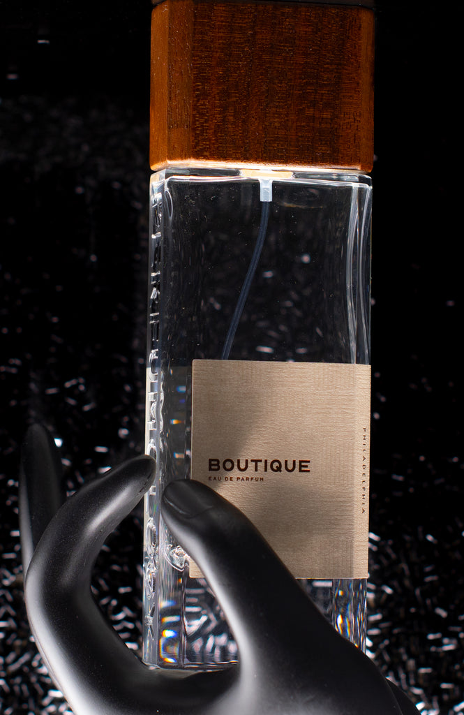 A clear glass bottle labeled "Perfumology Boutique" is held by a black mannequin hand. The perfume bottle, which has a wooden cap, is set against a speckled black background. Its essence combines fruity floral notes with undertones of woody white musk.