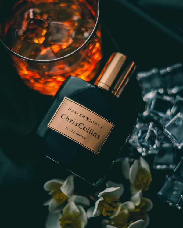 Positioned next to a glass of dark, ice-filled liquid, some loose ice cubes, and delicate white flowers lies a bottle of Chris Collins' "Harlem Nights" eau de parfum—an evening fragrance that whispers seduction.