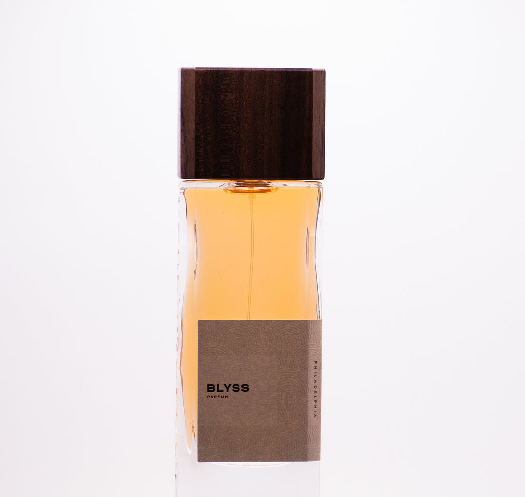 A clear glass perfume bottle with a wooden cap and a light brown label displaying the text "Blyss." The bottle, crafted with high quality ingredients, contains amber-colored liquid that embodies the essence of Perfumology fragrance.