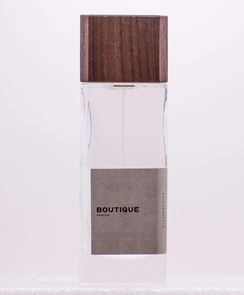 A clear glass perfume bottle with a wooden cap and a beige label that reads "Boutique by Perfumology," exuding a delicate blend of fruity floral and woody white musk notes.
