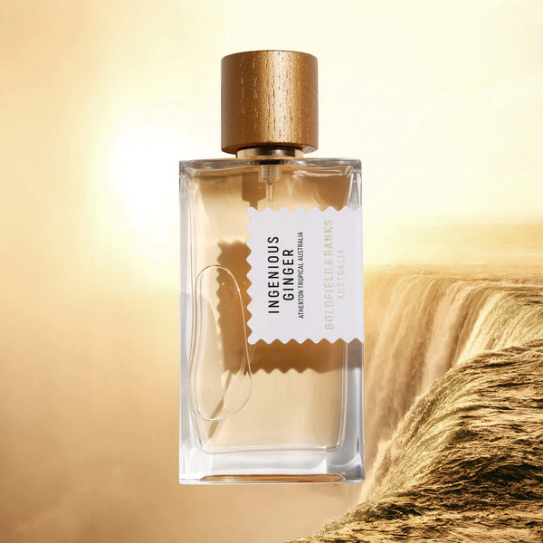 A transparent bottle of "Ingenious Ginger" perfume by Goldfield & Banks, with a gold cap, displayed against a backdrop of a flowing tropical waterfall.