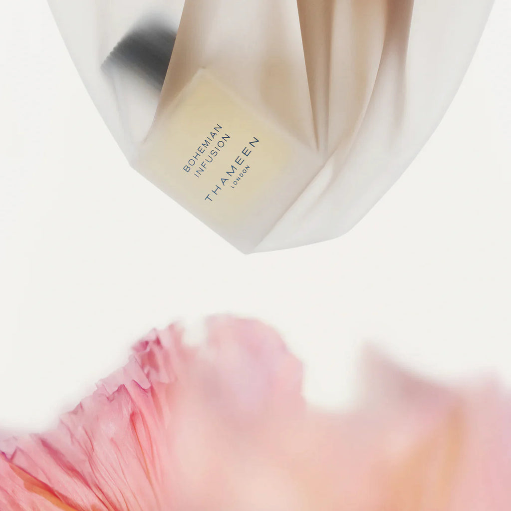 A bottle of THAMEEN Bohemian Infusion perfume is draped in a white fabric, with the image's lower foreground featuring blurred pink petals, highlighting the fragrance contrasts expertly crafted by Maurice Roucel.