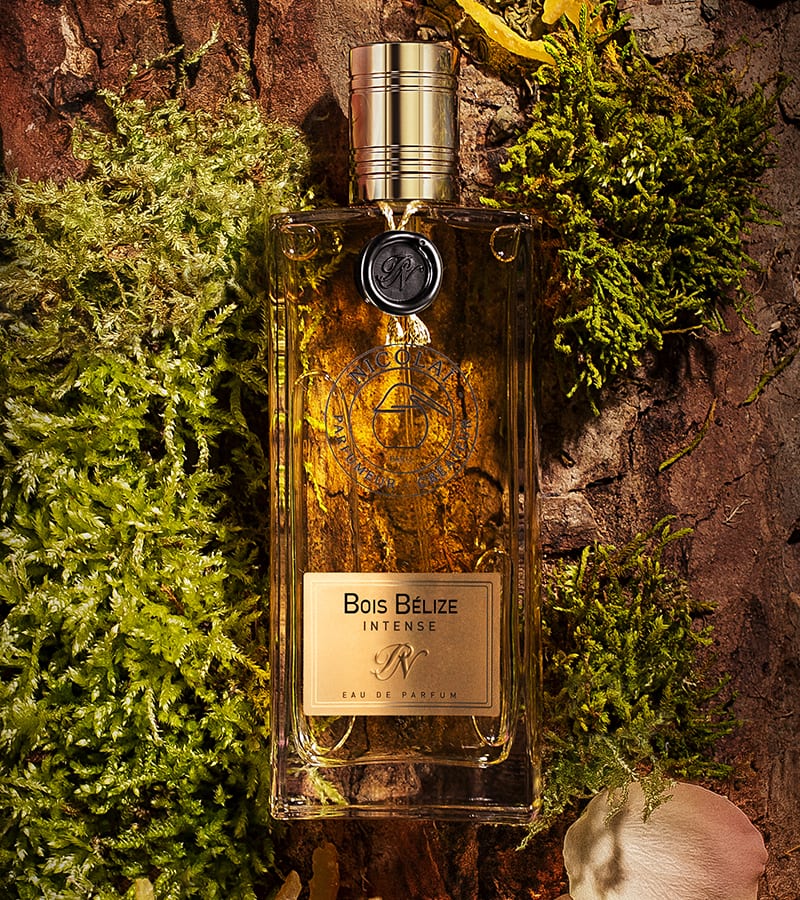 A bottle of "Bois Bélize Intense" Eau de Parfum by Nicolaï, with a gold cap, rests on a textured surface surrounded by green moss and natural elements, exuding subtle hints of guaiac wood.