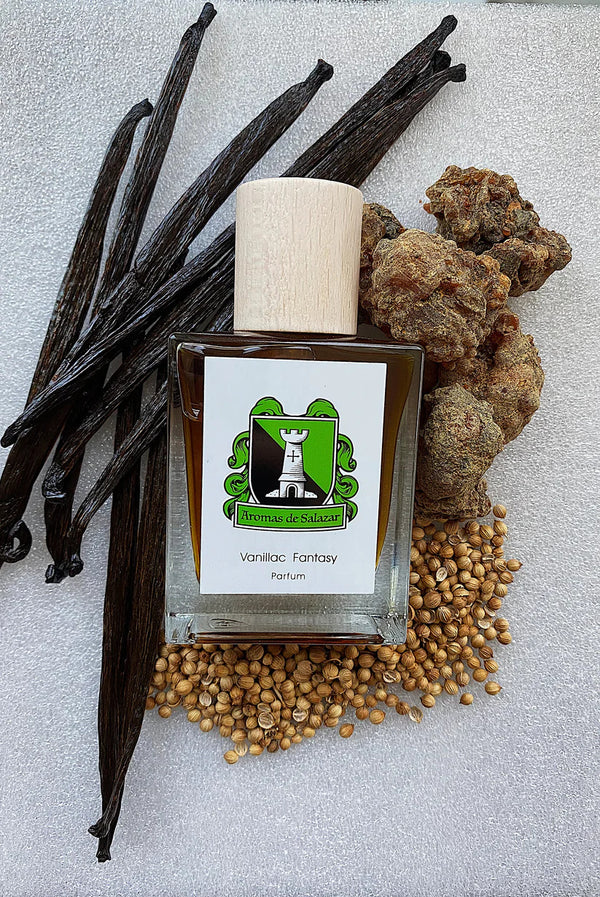 Bottle of Vanillac Fantasy by Aromas de Salazar, a unisex vanilla fragrance, is displayed with vanilla pods, seeds, and rocks on a textured white background.