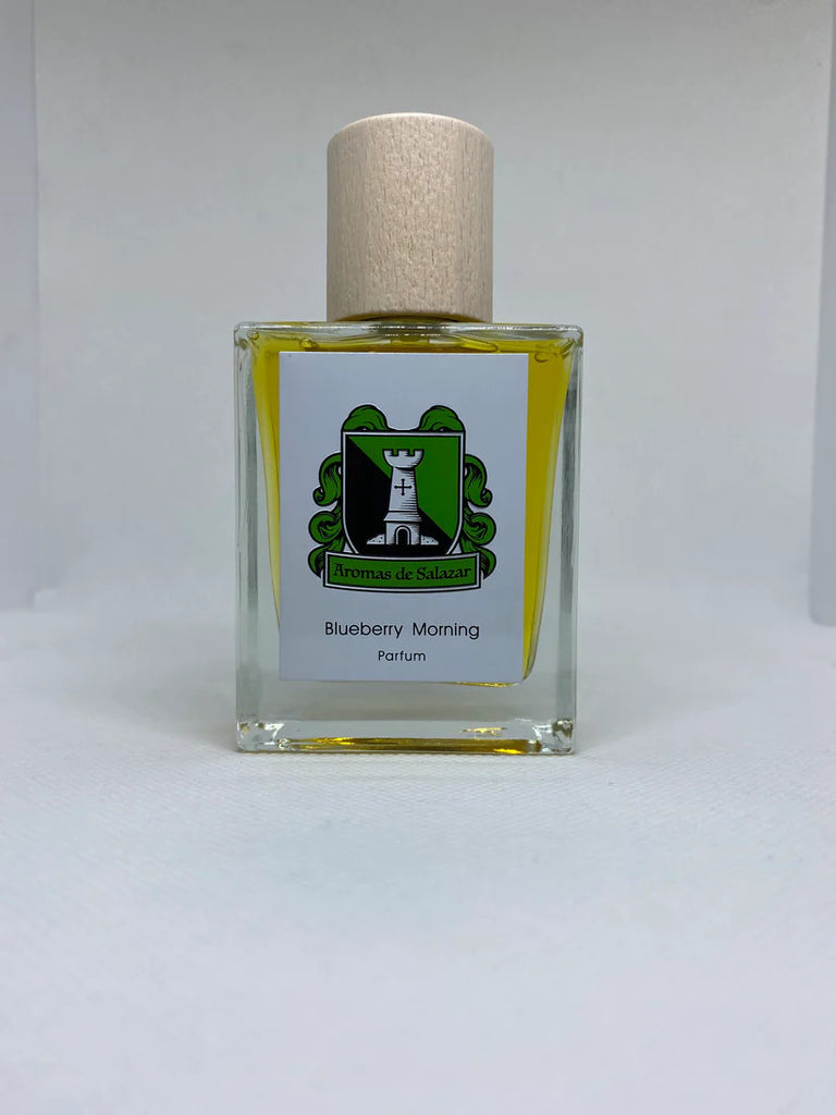 A clear glass bottle of perfume with a light wooden cap. The label reads "Aromas de Salazar, Blueberry Morning" and features a green and white emblem. Infused with the richness of Cambodian oud, this elegant bottle is placed on a neutral background.