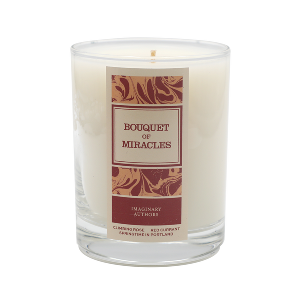 A white candle in a clear glass with a maroon and gold label that reads "Bouquet of Miracles" by Imaginary Authors. The label also mentions "Climbing Rose, Rose Red Currant, Springtime in Portland, Oregon.