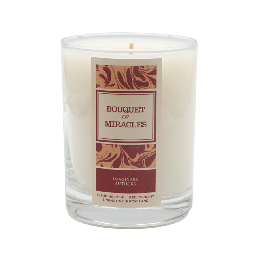 A white candle in a clear glass with a maroon and gold label that reads "Bouquet of Miracles" by Imaginary Authors. The label also mentions "Climbing Rose, Rose Red Currant, Springtime in Portland, Oregon.