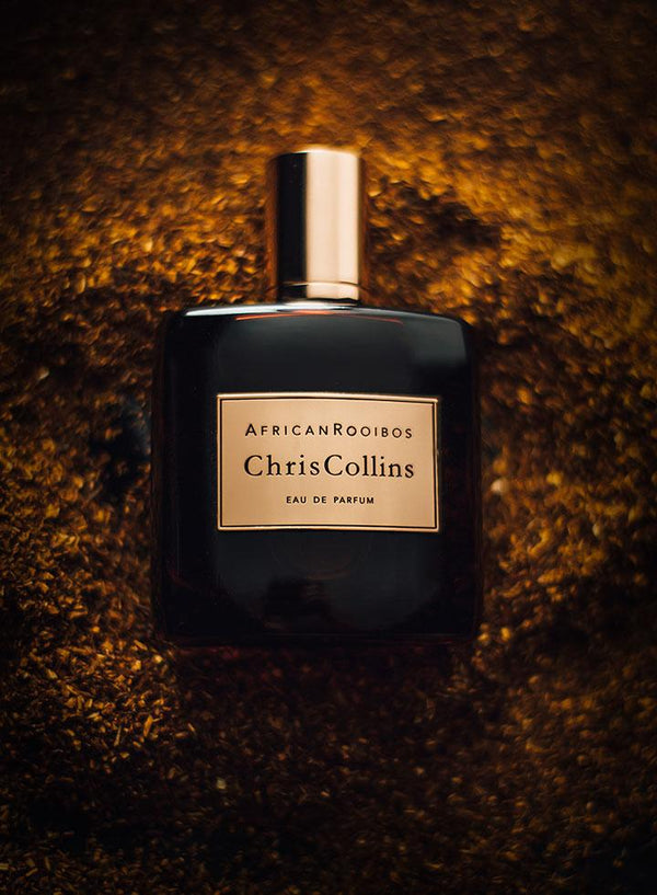 A bottle of Chris Collins African Rooibos is placed on a textured brown background. The dark bottle, adorned with a golden cap and label, evokes the rich essence of rooibos tea and cedarwood.