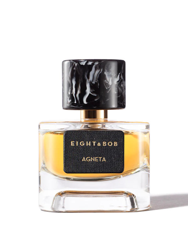 Transparent perfume bottle with a gold liquid inside, labeled "Eight & Bob Agneta," featuring a black marbled cap and a black rectangular nameplate from the Les Extraits Collection. This enigmatic fragrance is both alluring and sophisticated.