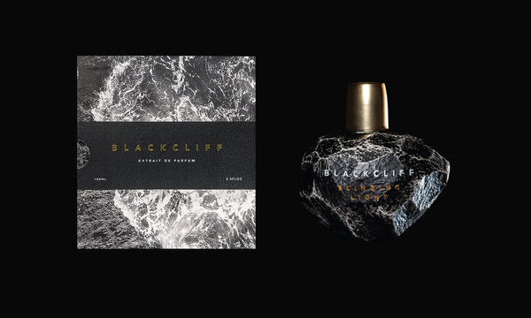 A black perfume bottle labeled "Blinding Light" by Blackcliff sits next to its box, which features a dark, wave-like design. Created by Kyle Mott-Kannenberg, the text on both the bottle and box states "Extrait de Parfum," showcasing an aromatic contradiction that captivates the senses.