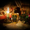 A lit candle, a bottle labeled "Zoologist: Bat," an inkwell with a feather quill, crystals, and rolled parchments are arranged on a table. The scent of Zoologist: Bat perfume lingers in the air. Books are stacked in the background, reminiscent of a cavernous home filled with ancient knowledge.