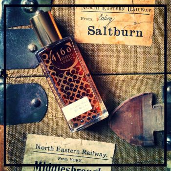 A bottle of Be Careful What You Wish For by 4160Tuesdays lies on a fabric surface adorned with vintage North Eastern Railway labels from Saltburn and Middlesbrough, its essence hinting at rich patchouli and guaicwood.
