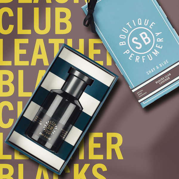 Image of a black perfume bottle labeled "Black Club Leather" by Shay and Blue in an open box with a blue and white striped interior. An unopened blue box with the same branding lies next to it on a brown surface with yellow text, highlighting this cruelty free fragrance.
