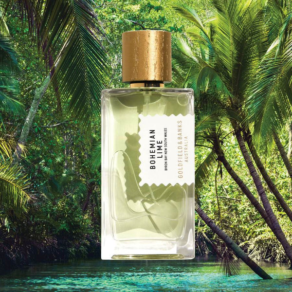 A glass bottle of "Bohemian Lime" fragrance by Goldfield & Banks is shown suspended against a lush, green forest background. The bottle, capturing the essence of Australian finger lime, features a gold cap and a white label.