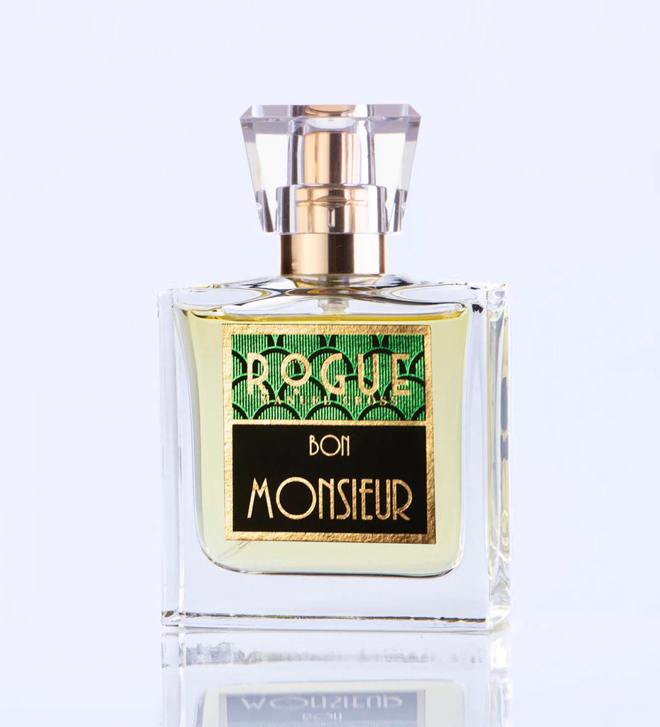 A glass bottle of "Bon Monsieur" by Rogue Perfumery with a gold cap and a green and black label sits on a white surface, exuding the classic charm of a gentlemen's fragrance with hints of fougeres and lavender.