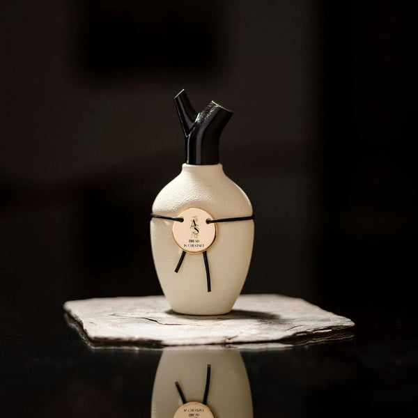 A white, vase-shaped perfume bottle with a black cap and a gold medallion sits on a dark, reflective surface, exuding an air of Bread in Chestnut by Scents of Wood touched with sandalwood.