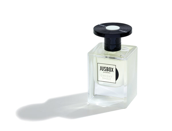 A transparent glass bottle of Jusbox's "Cheeky Smile" perfume, adorned with a sleek black cap, casts a gentle shadow on a pristine white background, radiating the vibrant energy of Acid House through its unique molecular fragrance.