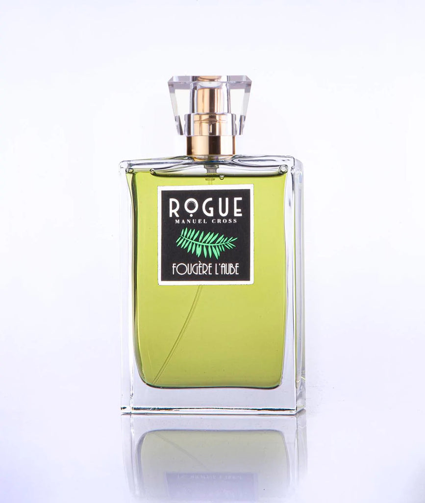 A clear glass bottle of Rogue Perfumery's "Fougere L'Aube" fragrance reveals a green citrus-infused, greenish-yellow liquid inside, topped with a gold-colored cap and a transparent cover.