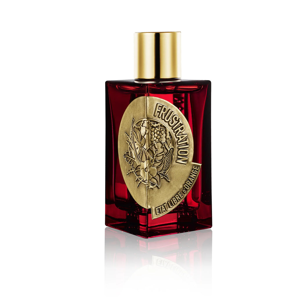 A red glass bottle with a gold cap containing a perfume named "Frustration EDP" by État Libre d'Orange. A gold emblem on the front depicts a detailed floral design, hinting at notes of vanilla and vetiver.
