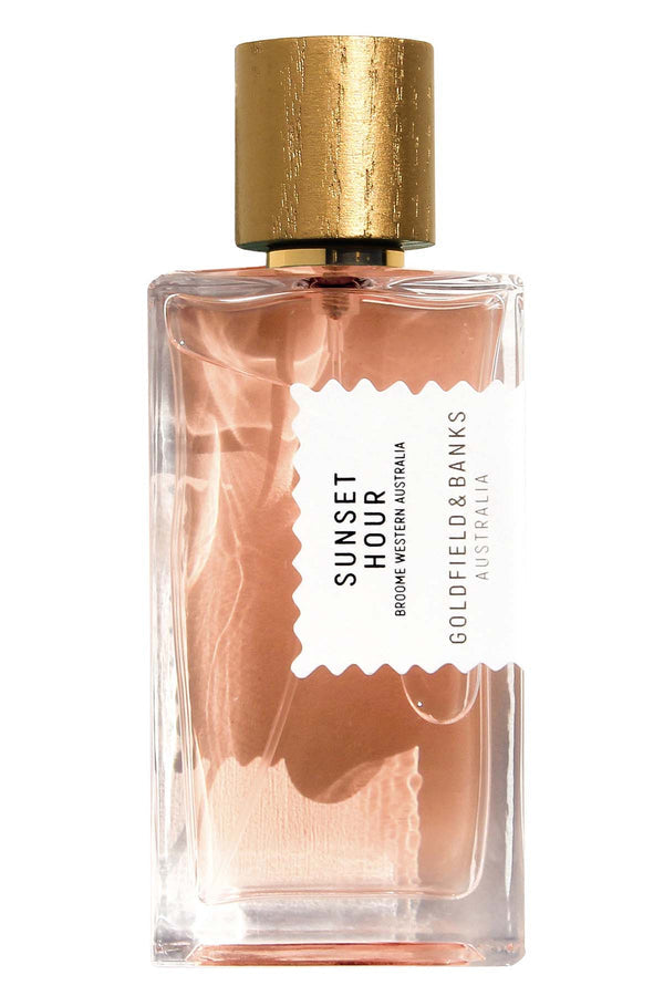 A clear glass bottle of Goldfield & Banks "Sunset Hour" perfume, featuring a golden cap and a white label with black text. The liquid inside is light pink, evoking the soft hues of Desert Peach.