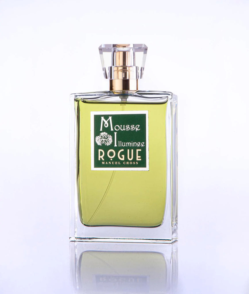 A clear rectangular perfume bottle with green liquid and a sophisticated label reading "Mousse Illuminee Rogue Perfumery," topped with a geometric design cap, exudes notes of Green Cypress.