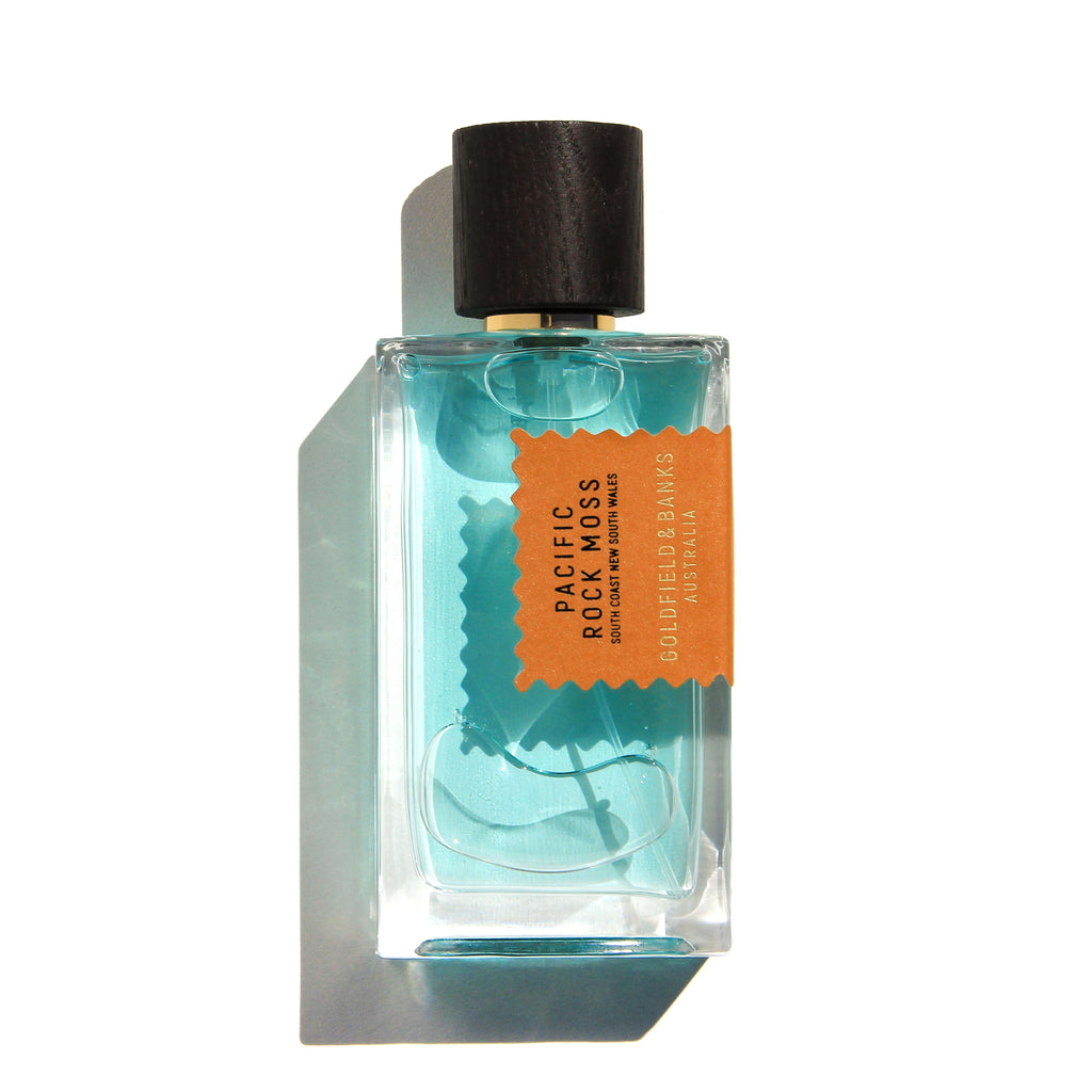 A bottle of Pacific Rock Moss by Goldfield & Banks, featuring light blue liquid and a dark wooden cap. The label is orange with gold text, perfectly capturing the essence of beach life in a fresh summer fragrance.