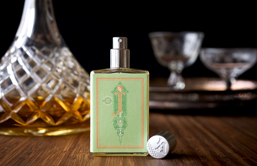 A bottle of Saint Julep by Imaginary Authors, featuring a decorative label and cap, sits on a wooden surface next to a cut glass decanter of bourbon with blurred glassware in the background.