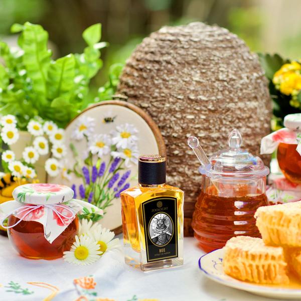 A collection of bee-themed items, including honey jars, a honeycomb, a beehive, and a Zoologist Bee perfume bottle with the rich aroma of honey, is arranged on a floral tablecloth with plants and flowers in the background.