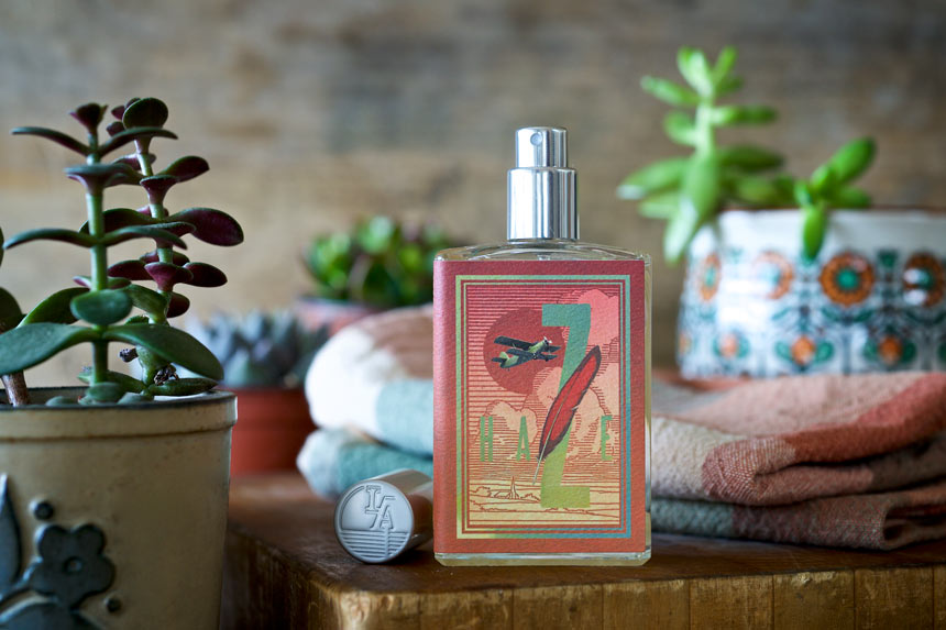 A bottle of Yesterday Haze by Imaginary Authors, with a seductive scent and decorative label, sits on a wooden surface amidst small potted plants and stacked towels.