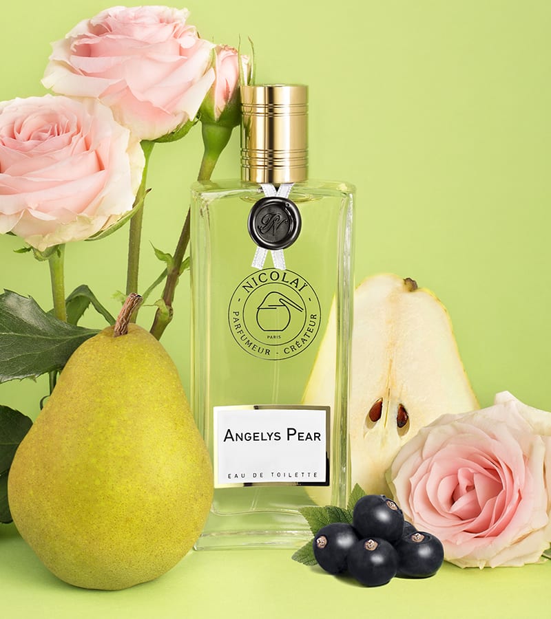 A clear bottle of Nicolaï Angelys Pear eau de toilette, embodying a fresh fragrance from the Loire Valley, is surrounded by a pear, blackcurrants, two whole roses, one cut rose, and a halved pear against a lime green background.