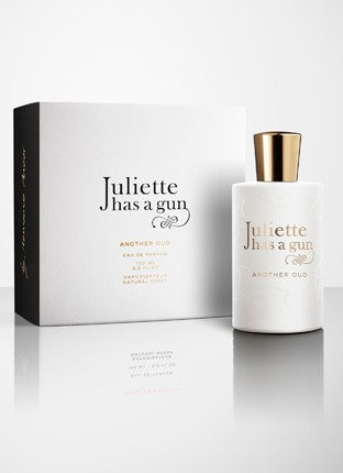 A white perfume bottle with a gold cap labeled "Juliette Has A Gun, Another Oud" sits elegantly beside its matching white box, boasting a woody composition and an exotic oriental fragrance that captivates the senses.