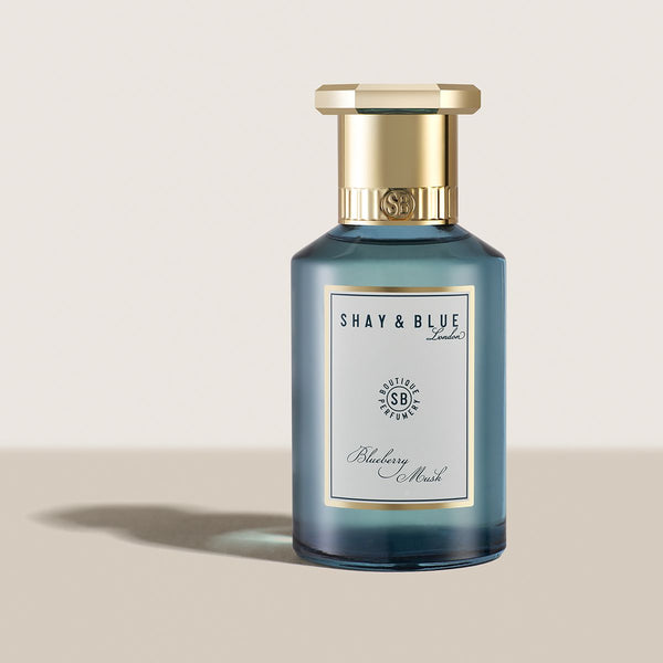 A bottle of Shay and Blue Blueberry Musk perfume with a blue body and gold cap, infused with hints of Orange Blossom, set against a beige background casting a shadow.