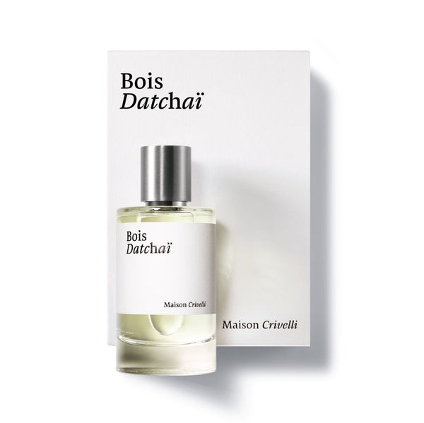 A bottle of Bois Datchai by Maison Crivelli is placed in front of its white packaging box. This woody fruity eau de parfum, enriched with black tea absolute, features a silver cap and clear glass bottle with a white label.