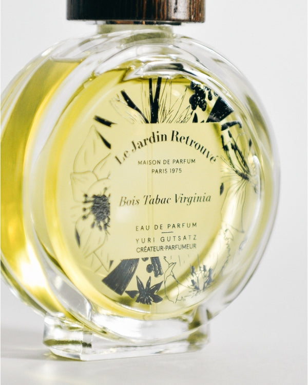 Clear bottle of 'Le Jardin Retrouvé' Eau de Parfum labeled "Bois Tabac Virginia Limited Edition" with yellow liquid inside and floral illustrations on the front, exuding a woody Eau de Parfum fragrance with hints of smoked vetiver.