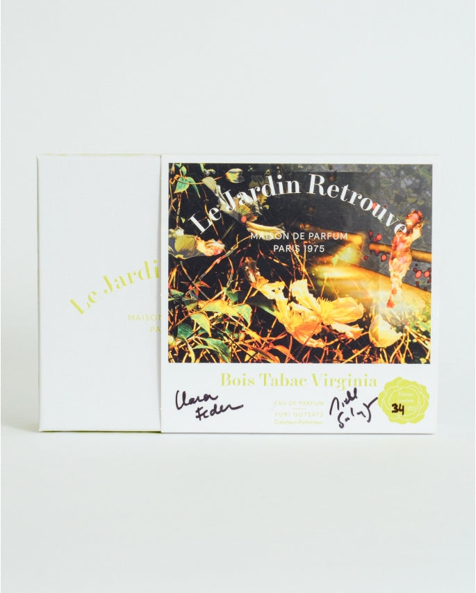 The image shows a white box with "Le Jardin Retrouvé" and "Bois Tabac Virginia Limited Edition" labels, hinting at the woody Eau de Parfum within. Signatures adorn the front, set against a leafy path where a person strolls.