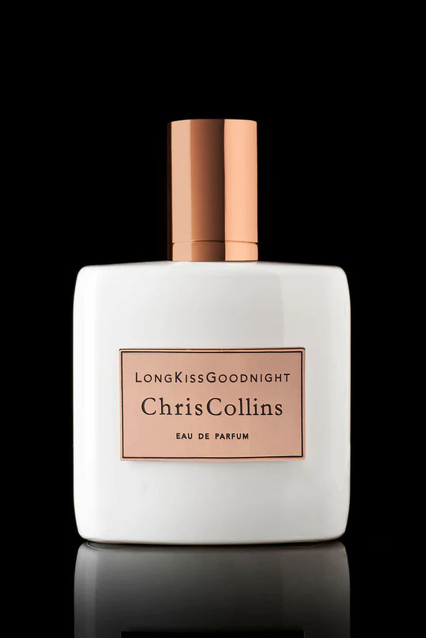 A white, rectangular perfume bottle with a rose gold cap features the label "Long Kiss Goodnight Eau de Parfum by Chris Collins" in black text on a rose gold background. The woody and floral fragrance stands out against a sleek black backdrop.