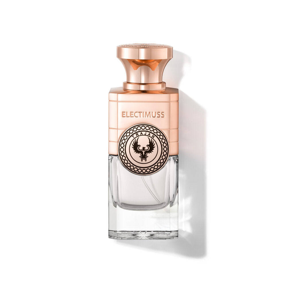 The perfume bottle called "Trajan" by Electimuss is rectangular in shape with a transparent lower half and features a rose-gold cap and top section. The label reads "ELECTIMUSS" above a circular emblem featuring a bird design, symbolizing an aromatic woody heart inspired by the Silk Route.