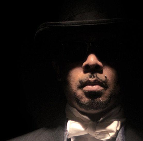 In a dimly lit scene, a man dons the LE DUC top hat, sunglasses, and bow tie by Ikiryo. His partially illuminated face exudes a masculine allure with an air of mystery.