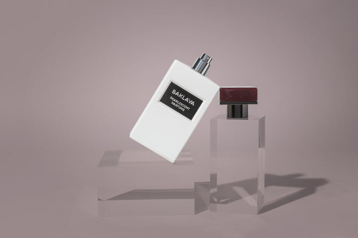 A white Pearlescent Parfums-branded Baklava bottle with a black label is displayed, slightly tilted, with a detached maroon cap nearby on two transparent stands against a plain background.