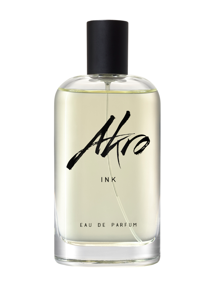 A clear glass bottle of INK by Akro, a unique tattoo fragrance with a black cap and black text on the label. The light yellow liquid inside captures the essence of body decoration.